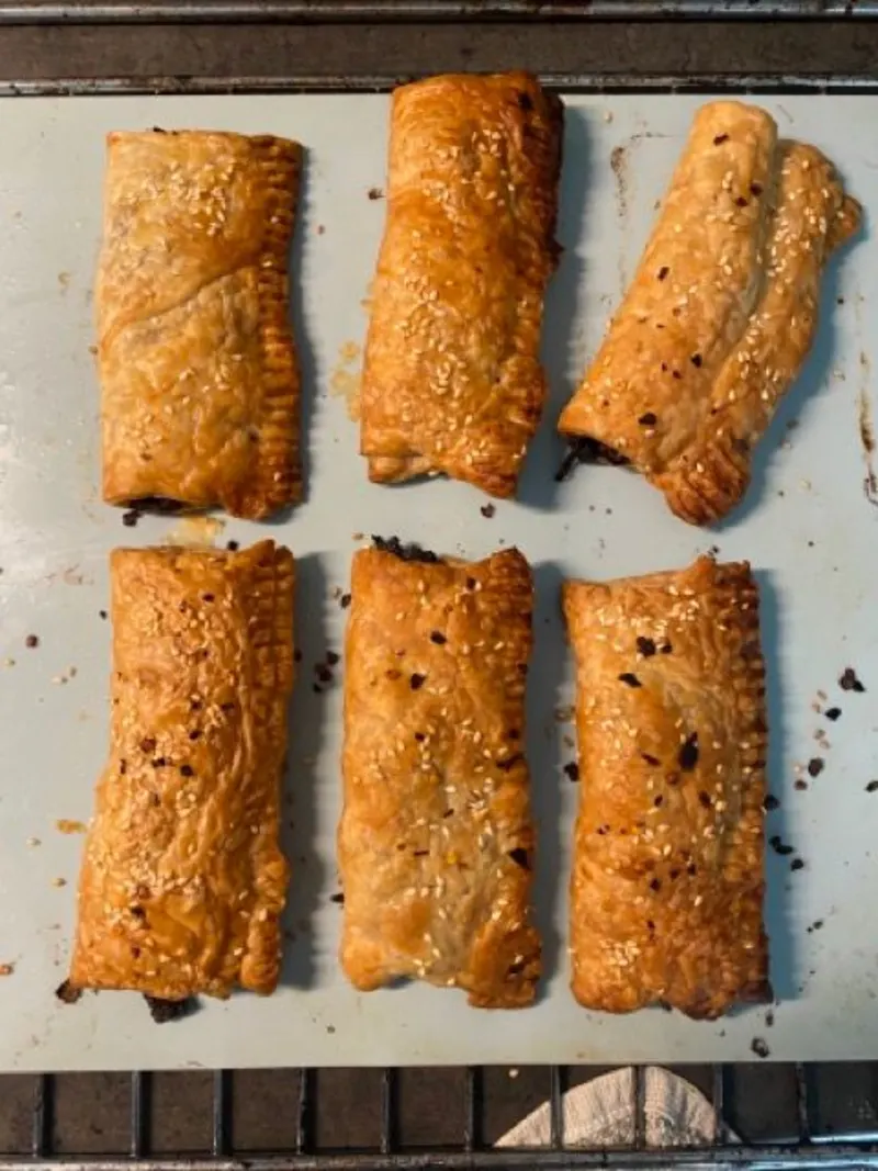 Een siliconen bakvel met zes saucijzenbroodjes liggen. Drie boven en drie onder. Het saucijzenbroodje rechtsboven ligt een beetje speels schuin. 