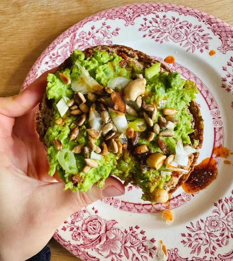 Een hand houdt een broodje vast met erwtendip met erop zonnebloempitten en cashewnoten