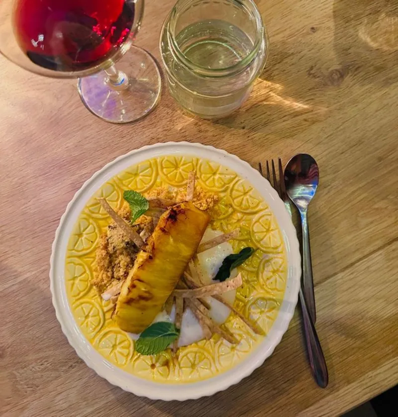 Een geel bord met grapefruit custard, gegrilde ananas, maisblad en een glas rode wijn