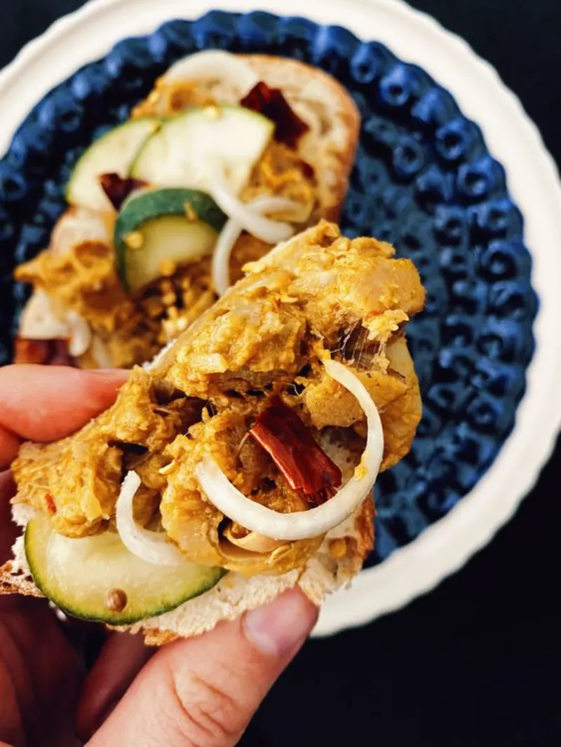Een etoaste boterham op een blauw bord. Op deze boterham zie je rendang van jackfruit. Een stukje brood wordt er boven vast gehouden door twee vingers.