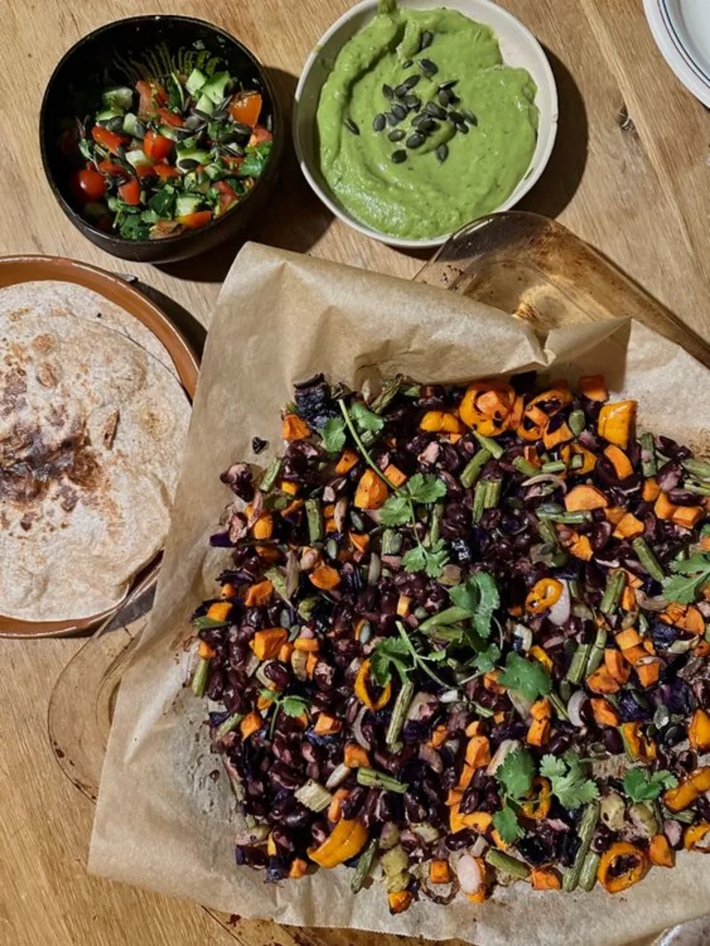 Op een tafel, een bordje met tortillas, een ovenschaal met geroosterde groenten, een bakje salsa en avocadoroom