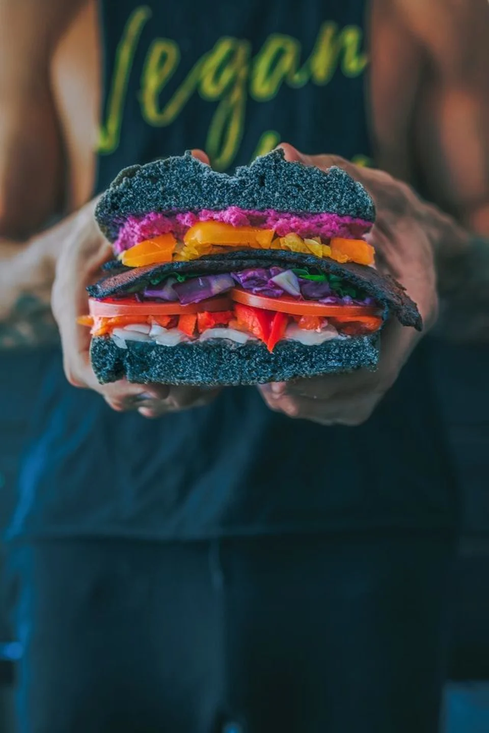Twee handen die een zwart burger broodje met allerlei gekleurde ingredienten erop vast houden. 