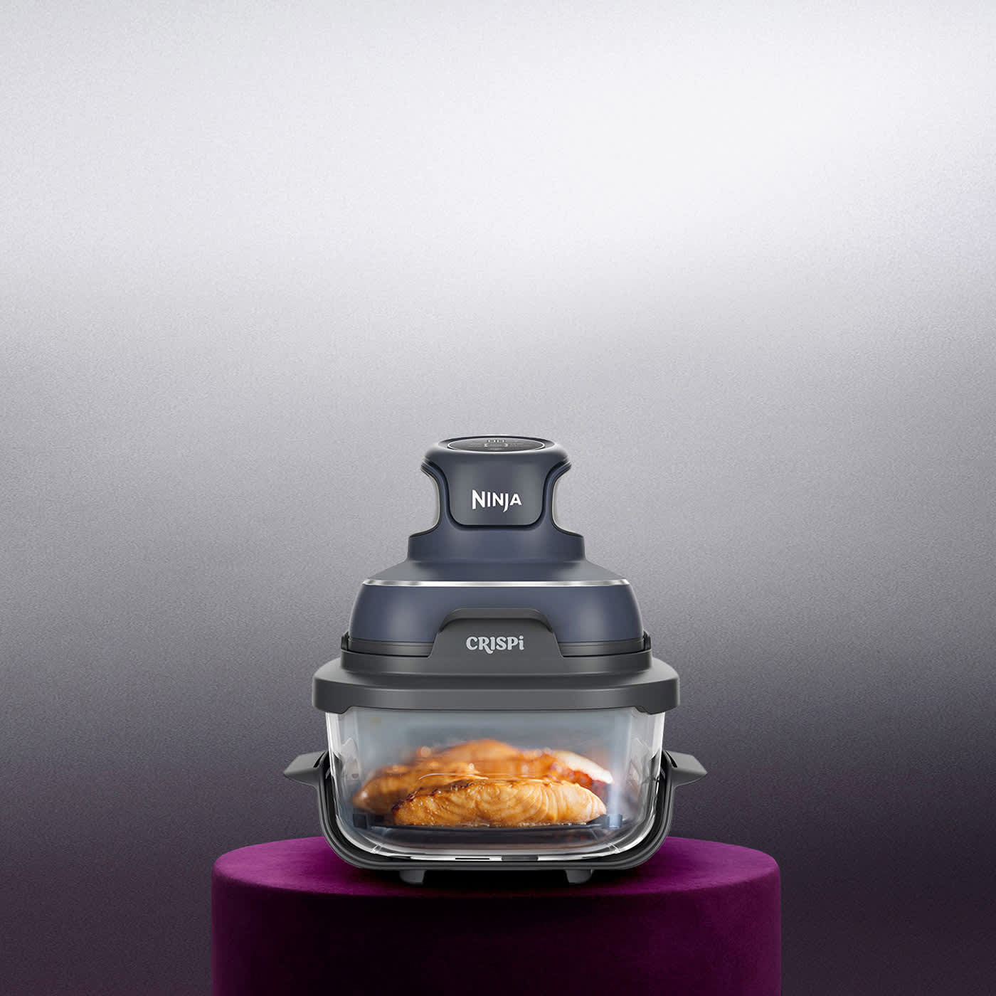 Image of Ninja Crispi 4-in-1 portable glass air fryer with food inside, displayed on a purple pedestal against a grey background.