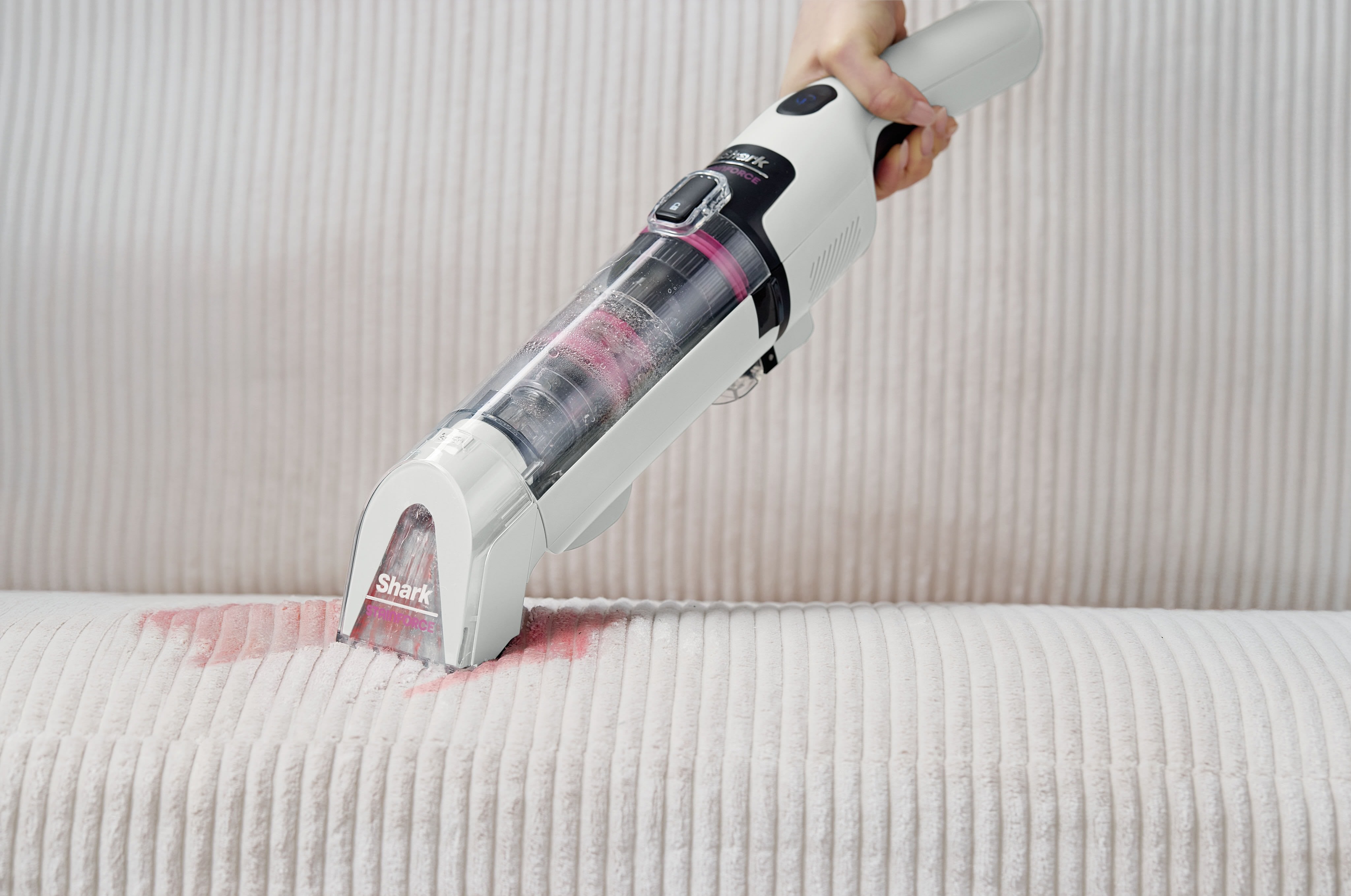 Une personne utilise un aspirateur à main sans fil Shark StainForce pour nettoyer du vin rouge renversé sur une surface de rembourrage de couleur claire.