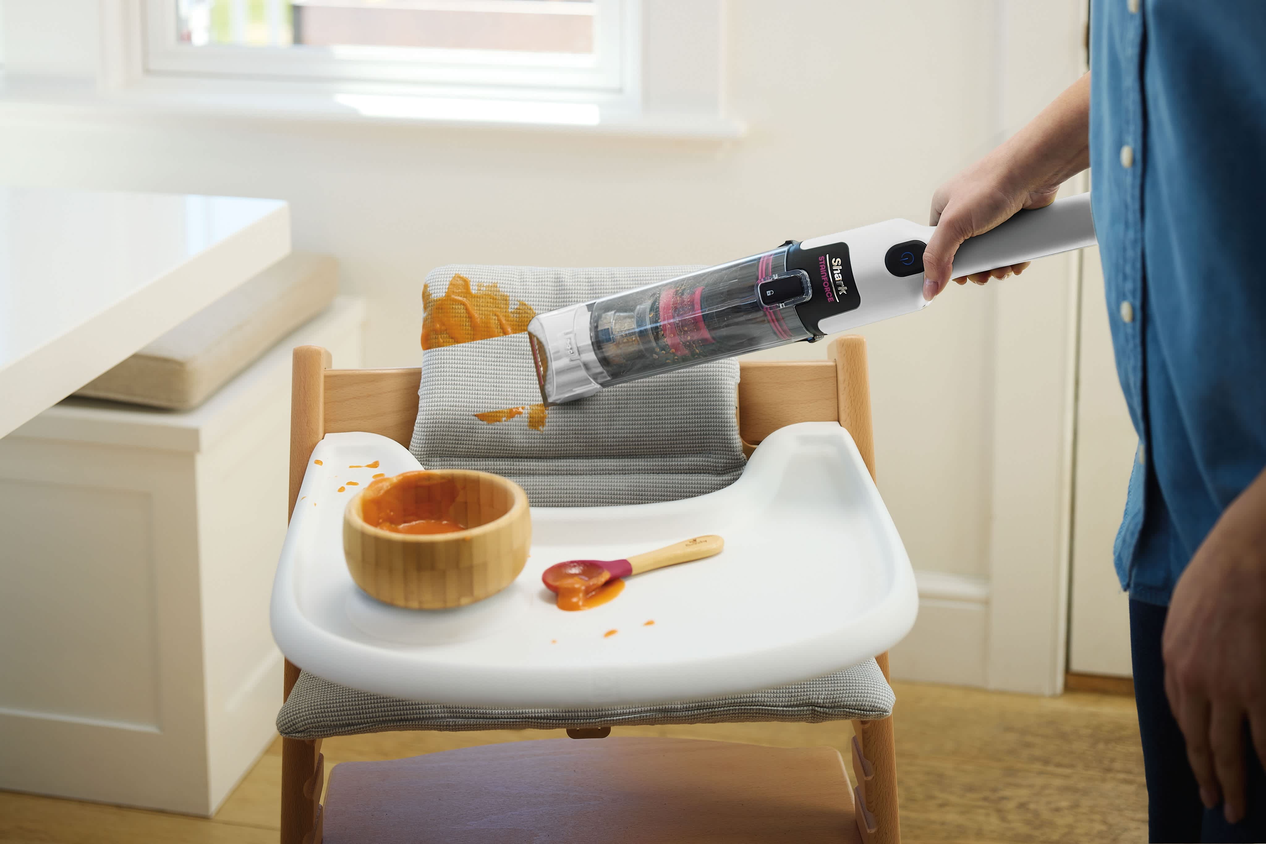 Une personne utilise un aspirateur à main sans fil Shark StainForce pour nettoyer les aliments pour bébé renversés sur le plateau et le coussin d'une chaise haute.