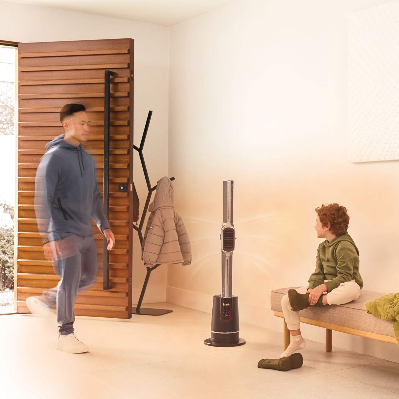 A man enters a bright room as a boy sits on the bench putting on shoes, with a Shark Turbo Blade cool and heat fan