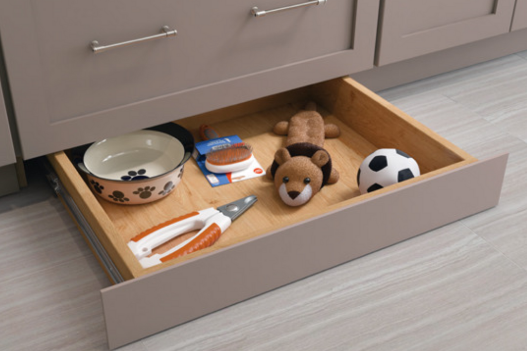 A grey and natural wood toe kick drawer filled with pet toys, pet bowl, and pet grooming tools.