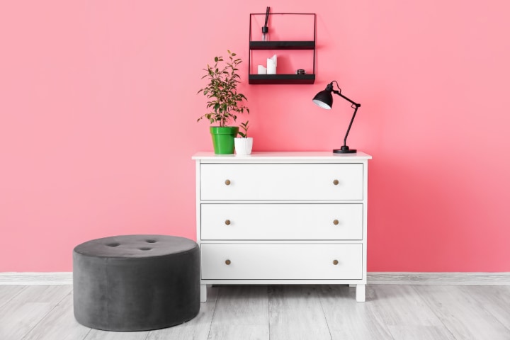 White chest of drawers and black ottoman in front of a pink wall