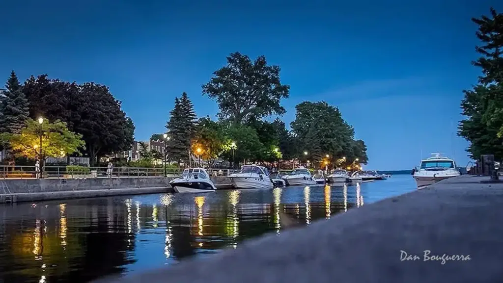 Boats docked along a peaceful canal at dusk in West Island, Montreal, with trees and lampposts reflecting in the water under a deep blue evening sky.