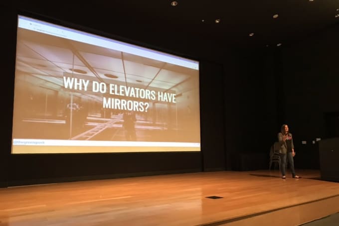 Sia speaking on a large stage with a slide showing 'Why do elevators have mirrors?'