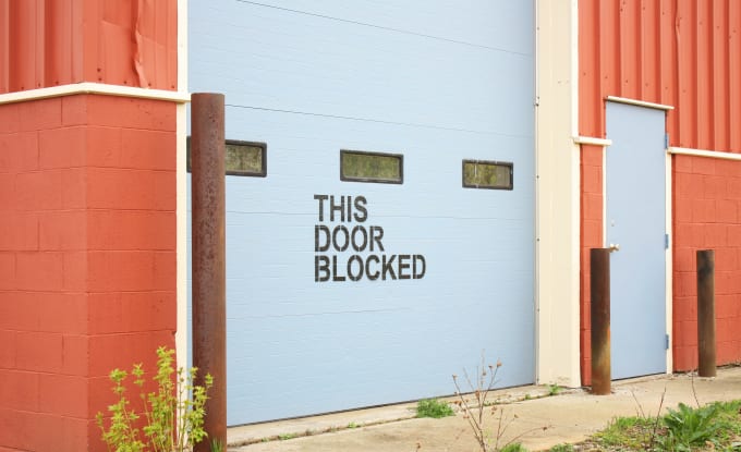 A white garage door with large, black, block lettering saying 'THIS DOOR BLOCKED'. The building is a bright orange color so the contrast is high.