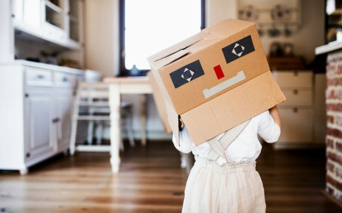 Toddler girl with an upside down cardboard box over her head. The box is decorated like a robot.