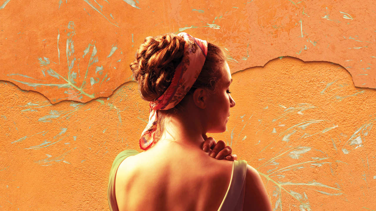 Woman with hair in a braided bun and floral headband facing towards a wall that's textured orange wall with paint splatters in leaf-like patterns