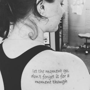 back of a woman's shoulder with a tattoo of the words let the moment go, don't forget it for a moment though