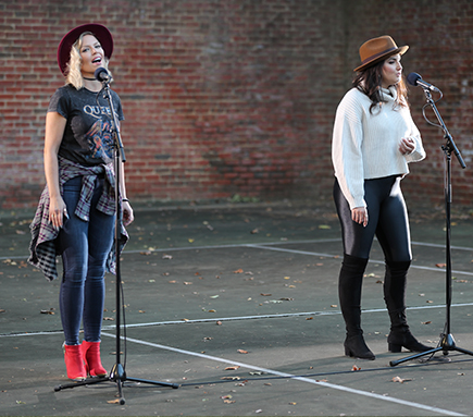 Crystal Mosser and Maria Rizzo singing at microphone stands 6 feet apart in front of a brick background