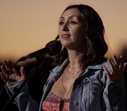 Natascia Diaz singing with arms raised in front of a microphone at sunset