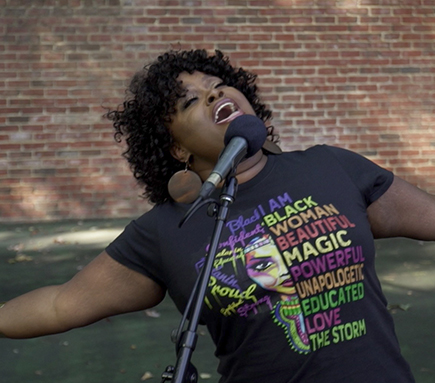 Nova Y. Payton exuberantly singing at a microphone stand in front of a brick wall