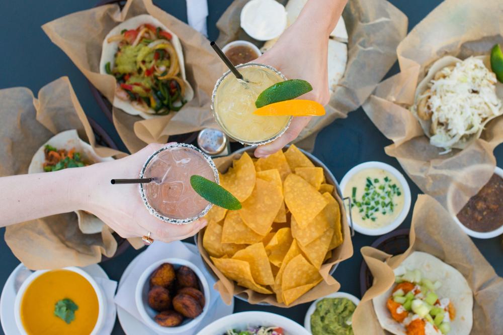 People tasting margaritas over tacos and chips and dip.