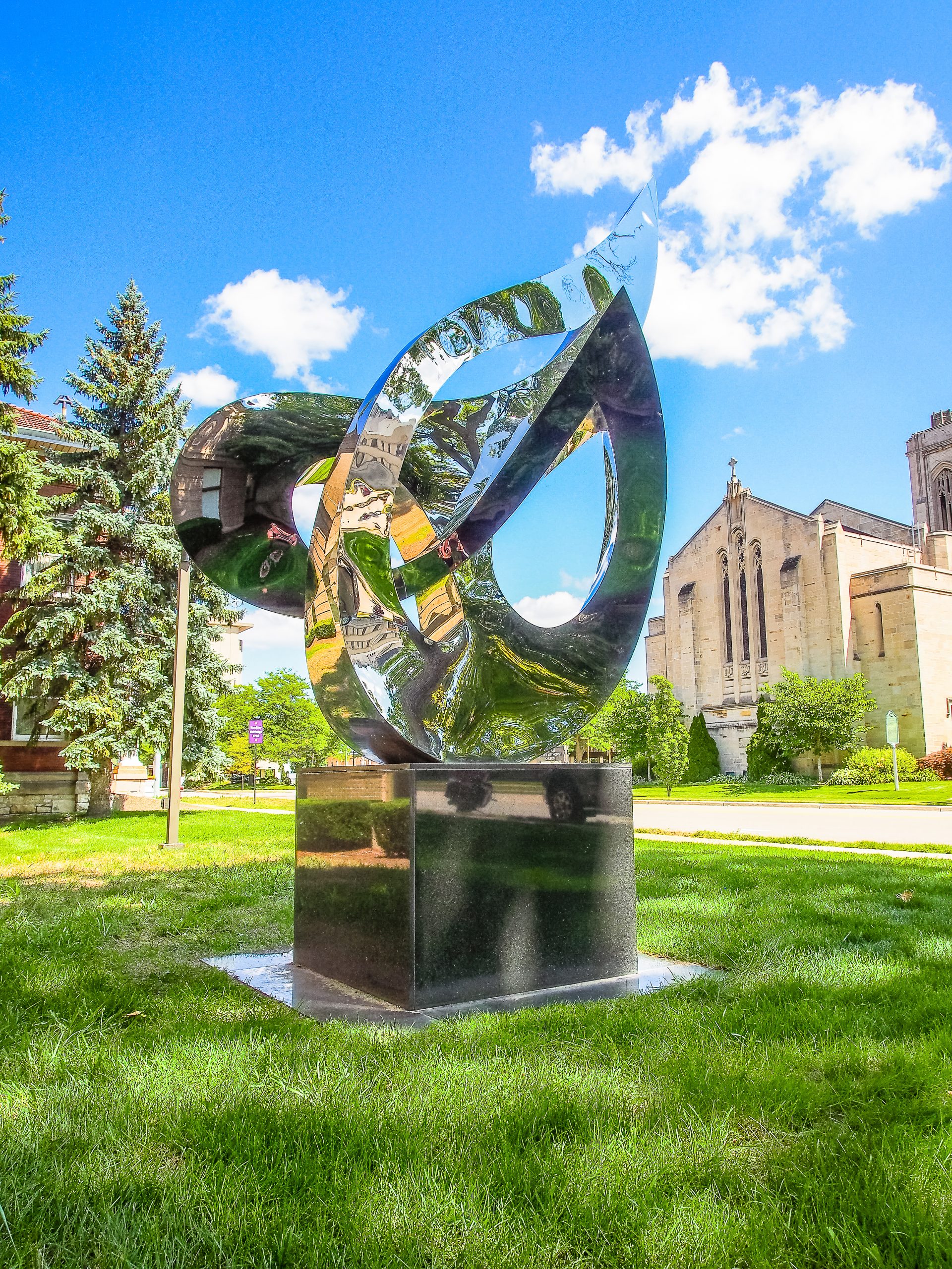 shiny, silver modern art sculpture sits on base. Green grass, blue sky and white clouds. Stone church sits to right, trees to left. 