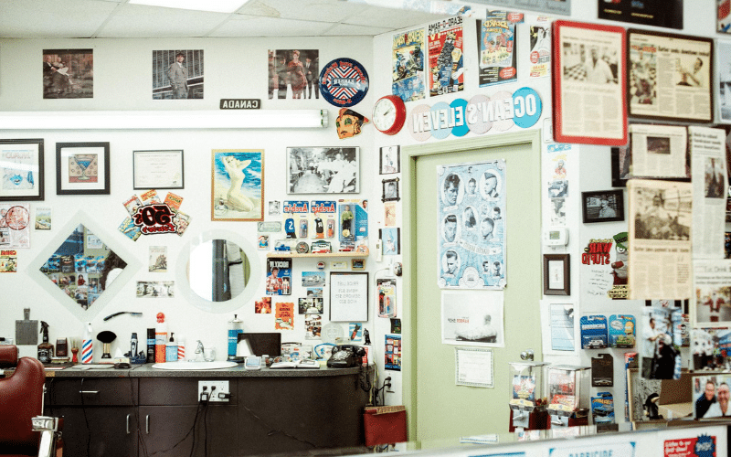 The interior of Dave's barbershop in Nanaimo, showing walls covered floor-to-ceiling with framed memorabilia, historical newspaper clippings, classic pin-up art, and a poster of vintage hairstyles. A red barber chair is visible in the foreground