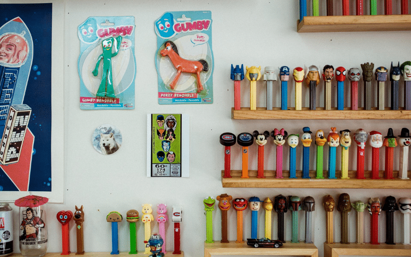 A close-up view of the Pez dispenser collection and other vintage pop-culture collectables displayed on wooden shelves inside Dave’s shop. Items include Gumby and Pokey figures and various character dispensers