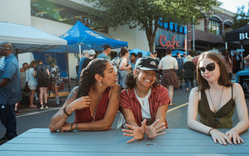 Photo of three women sitting together at an outdoor table during a street festival. On the left is Sonia Malek-Komen, sticking her tongue out at the woman in the centre, her daughter, who is wearing a brown baseball cap, a striped shirt, and a bright smile. On the right, a third woman with long brown hair and dark sunglasses is looking directly at the camera. The background is busy with market tents, crowds, and a storefront that reads "Cafe."