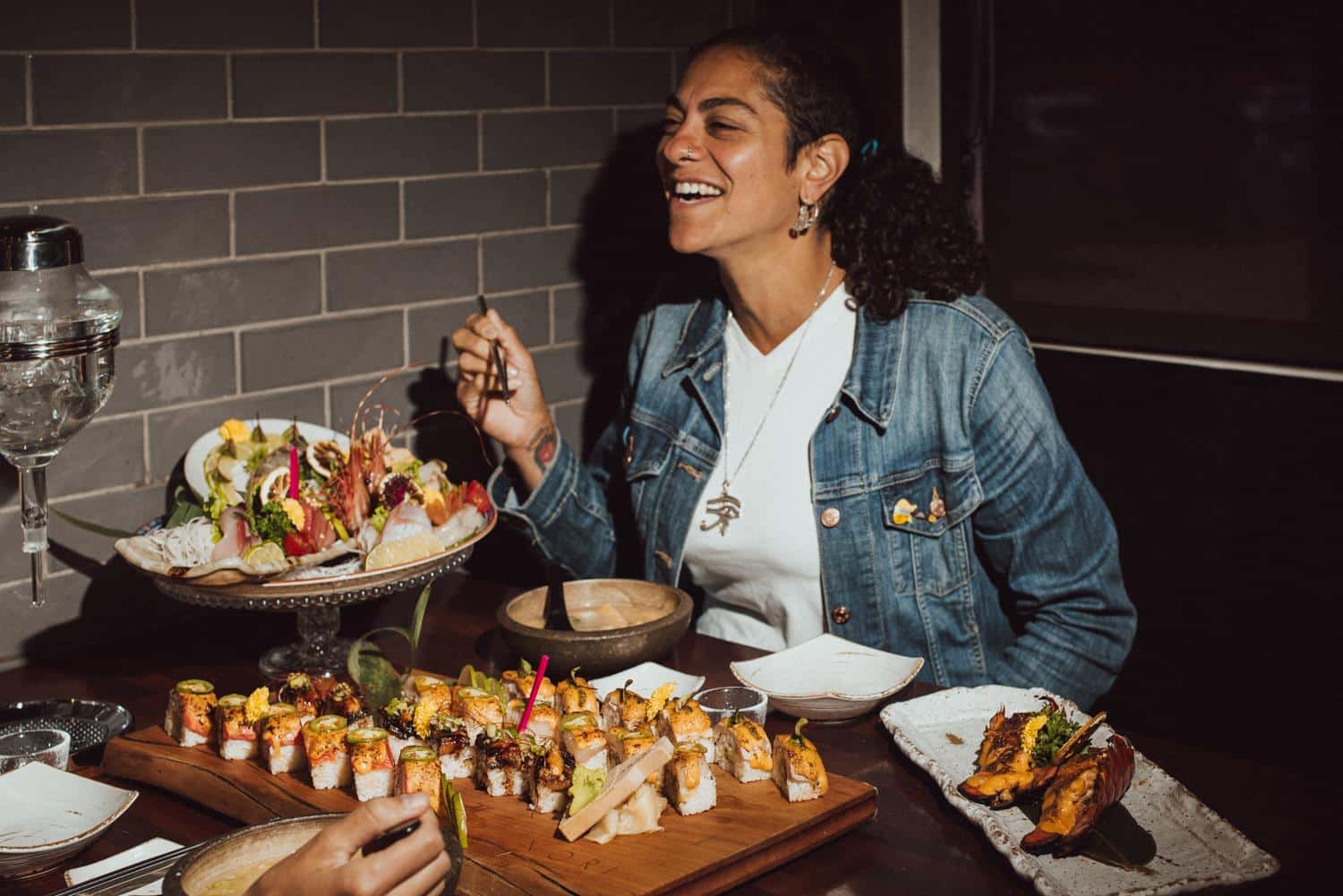 Photo of Sonia Malek-Komen seated at a restaurant table, laughing while holding chopsticks. She has her dark curly hair pulled back and is wearing a denim jacket and a white shirt. The table is laden with elaborate plates of sushi and sashimi, including rolls arranged on a wooden board and a large plate of sashimi salad on a tiered glass stand.
