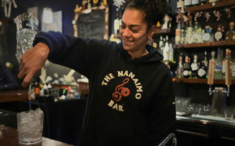 Photo of Sonia Malek-Komen, a woman with dark, curly hair and medium skin tone, smiling and standing behind a bar, while pouring a drink on the counter. She is wearing a black hoodie with 'The Nanaimo Bar' logo, and the background features well-stocked liquor shelves and holiday decorations.