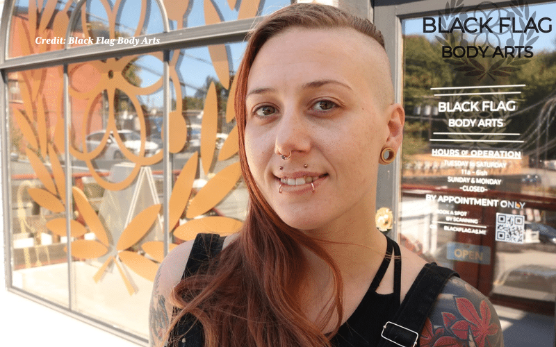 A light-skinned person with a shaved side head and long reddish-brown hair on the opposite side smiles slightly while standing in front of a storefront. They have multiple piercings, including double nose rings and a horizontal lip piercing. They are wearing a black tank top with visible shoulder tattoos and a large, wooden ear gauge. The reflection of stylized, orange leaf decals is visible on the window behind them, and the business name “BLACK FLAG BODY ARTS” is printed on the glass door.
Photo credit: Black Flag Body Arts