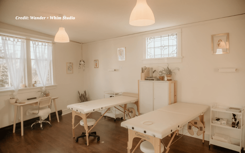 A picture of Wander + Whim’s bright, calm, and minimalist studio, bathed in warm, soft light. The walls are pale cream, and the floor is dark hardwood. The room contains two white tattoo tables with light wood legs. On the left, near a window with sheer white curtains, is a light wood desk with a laptop and a wicker-backed office chair. The room is decorated with small potted plants and framed artwork.
Photo credit: Wander + Whim Studio