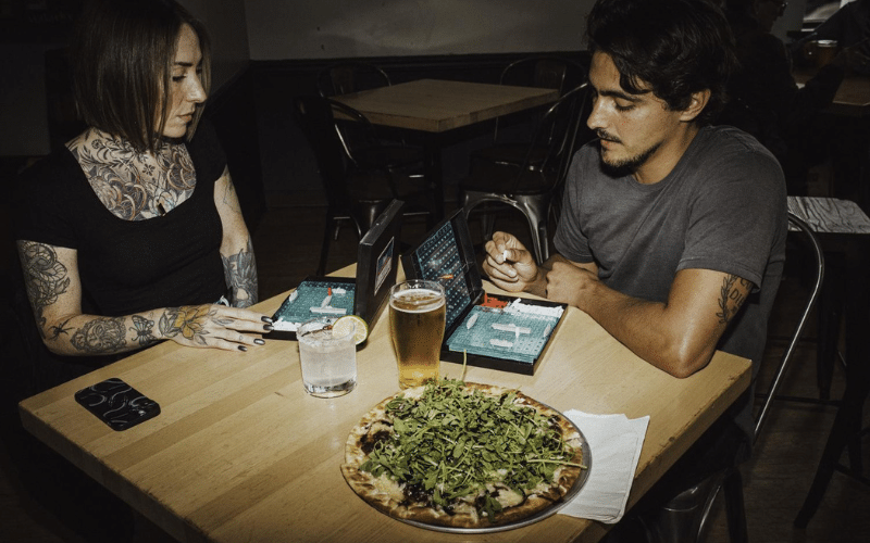 A photo of two people sit across from each other at a light wood table in a dimly lit, brewery setting. They are playing Battleship, with the blue game units open between them. One person, on the left, has shoulder-length dark hair and visible, intricate tattoos across their chest, neck, and arms. In the center of the table sits a large, thin-crust pizza topped with a mountain of fresh arugula, alongside a pint of beer and a glass of water with a lime wedge.