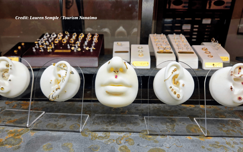 A close-up of a jewelry display on a slate surface. Five white anatomical silicone molds, four ears and one nose/mouth combo, are mounted on clear acrylic stands. They are decorated with an array of intricate piercings, including chains, hoops, and gem-encrusted studs. In the background, rows of rings and additional jewelry are visible on velvet and leather displays.
Photo credit: Lauren Semple