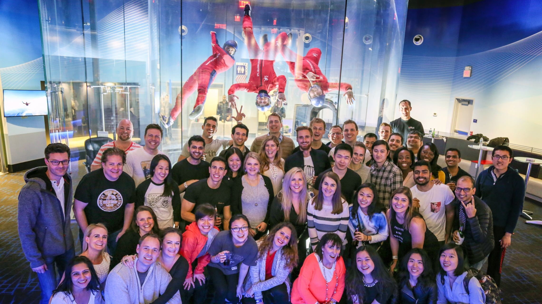 large group of people pose in front of wind tunnel as instructors are indoor skydiving