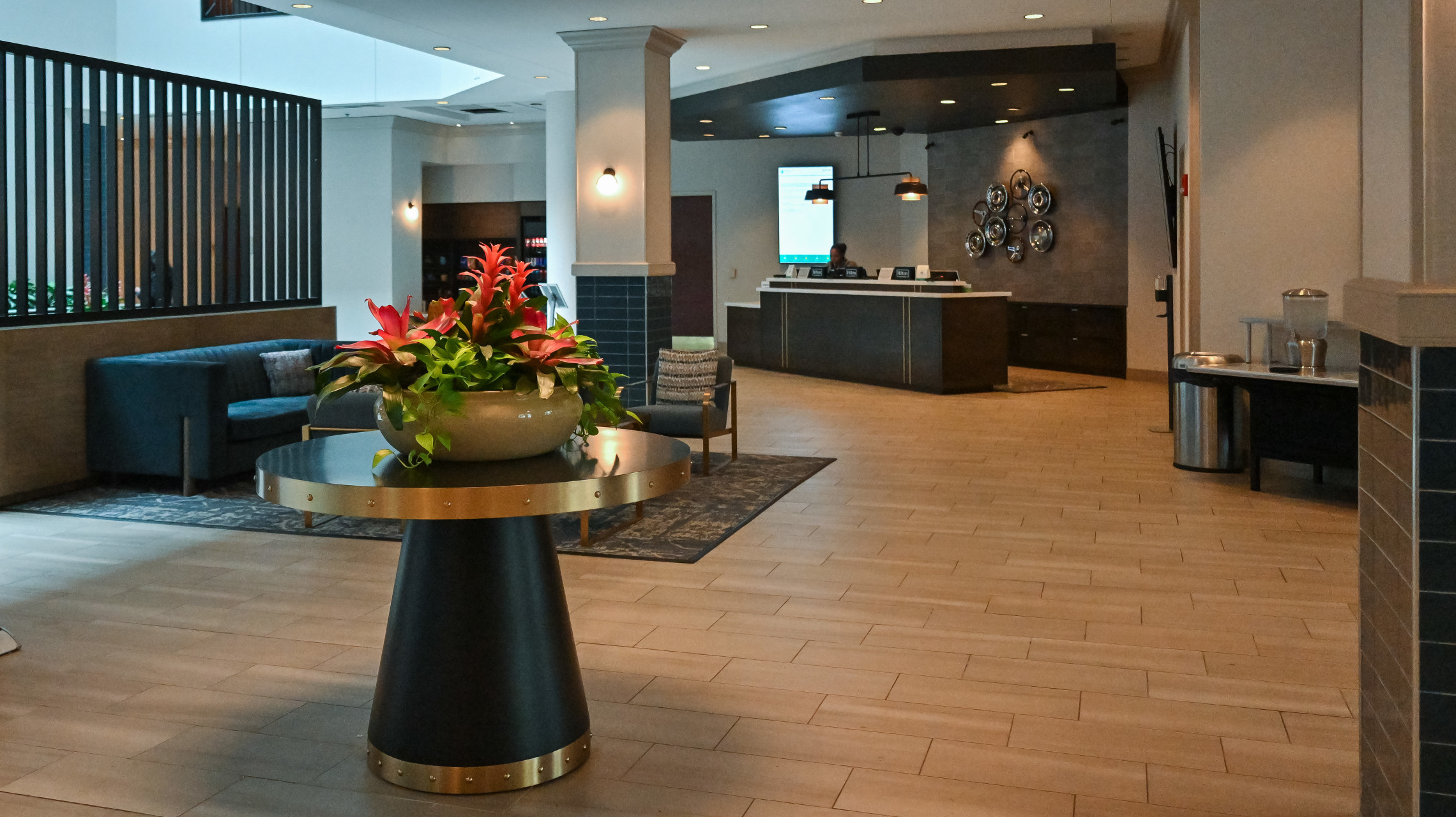 Photograph of a modern hotel lobby featuring a central round table with a large floral arrangement, seating areas with sofas, and a reception desk in the background. The space includes neutral tones, tiled flooring, decorative wall elements, and soft lighting creating a welcoming atmosphere.