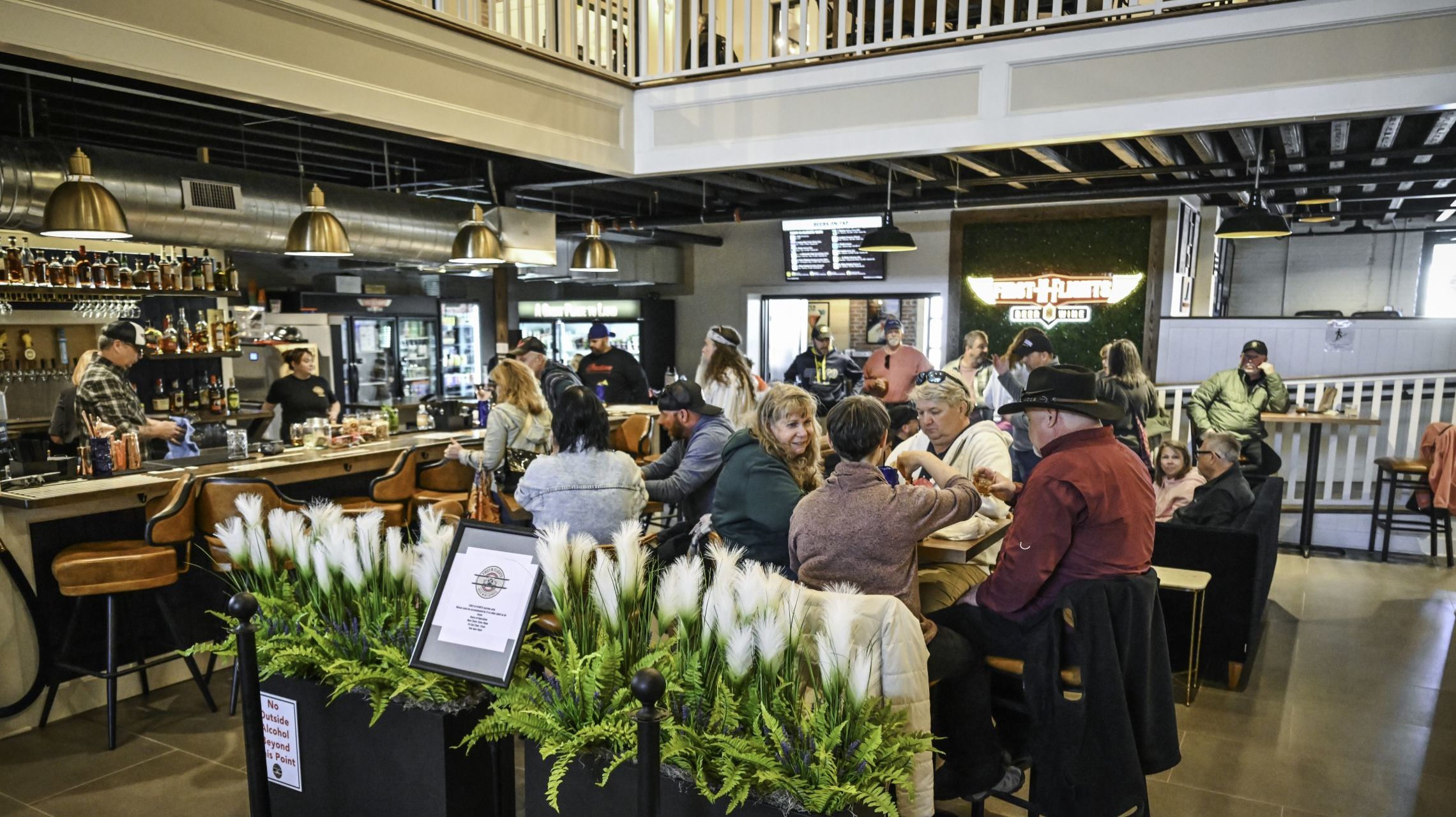 The image shows a busy, well-lit bar with people seated at various tables, enjoying drinks and socializing, surrounded by decorative plants and a modern, industrial design.