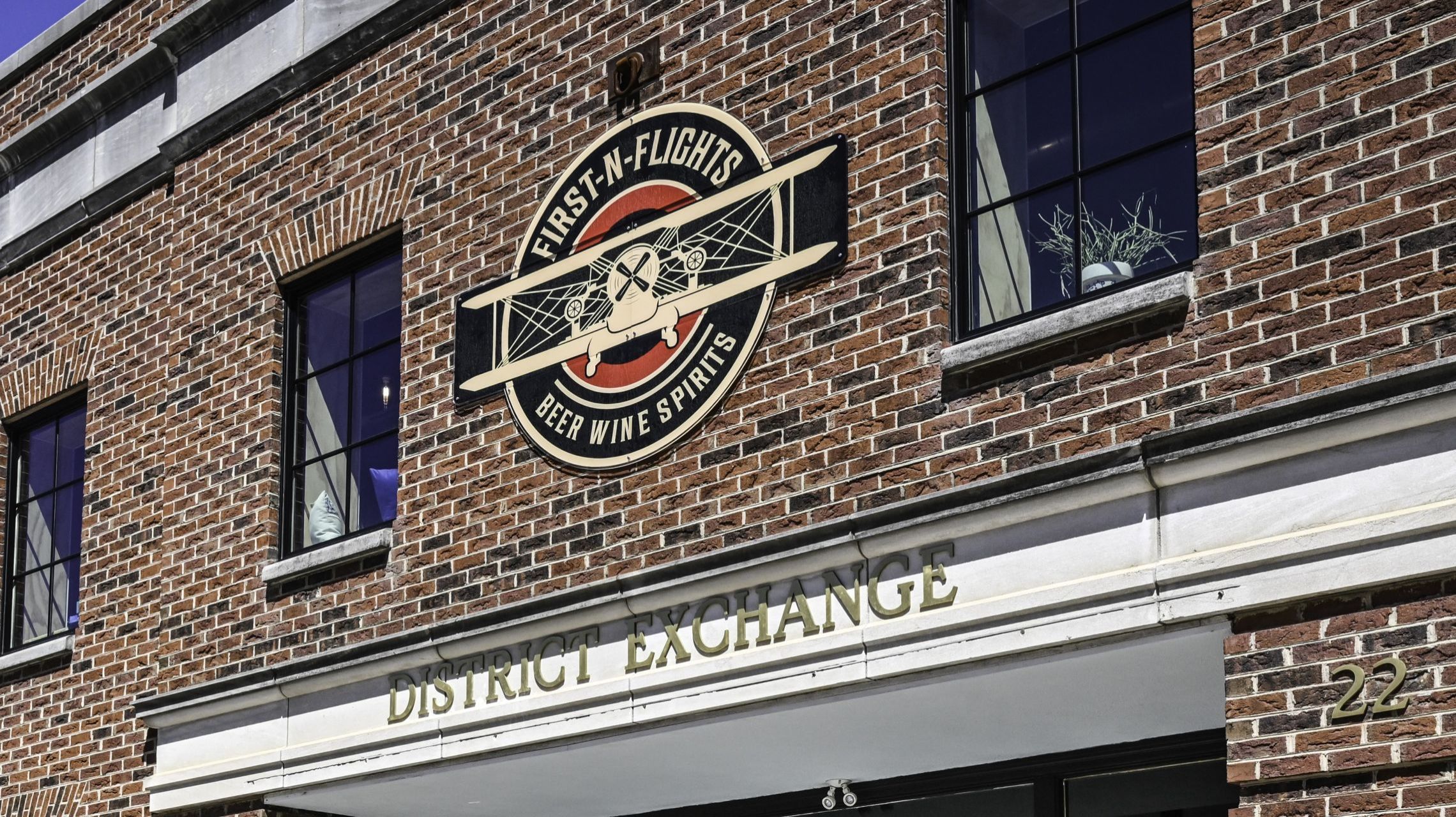 A brick building featuring the words "District Exchange" a sign with a propellor airplane reading "First -N- Flights Beer &amp; Wine"