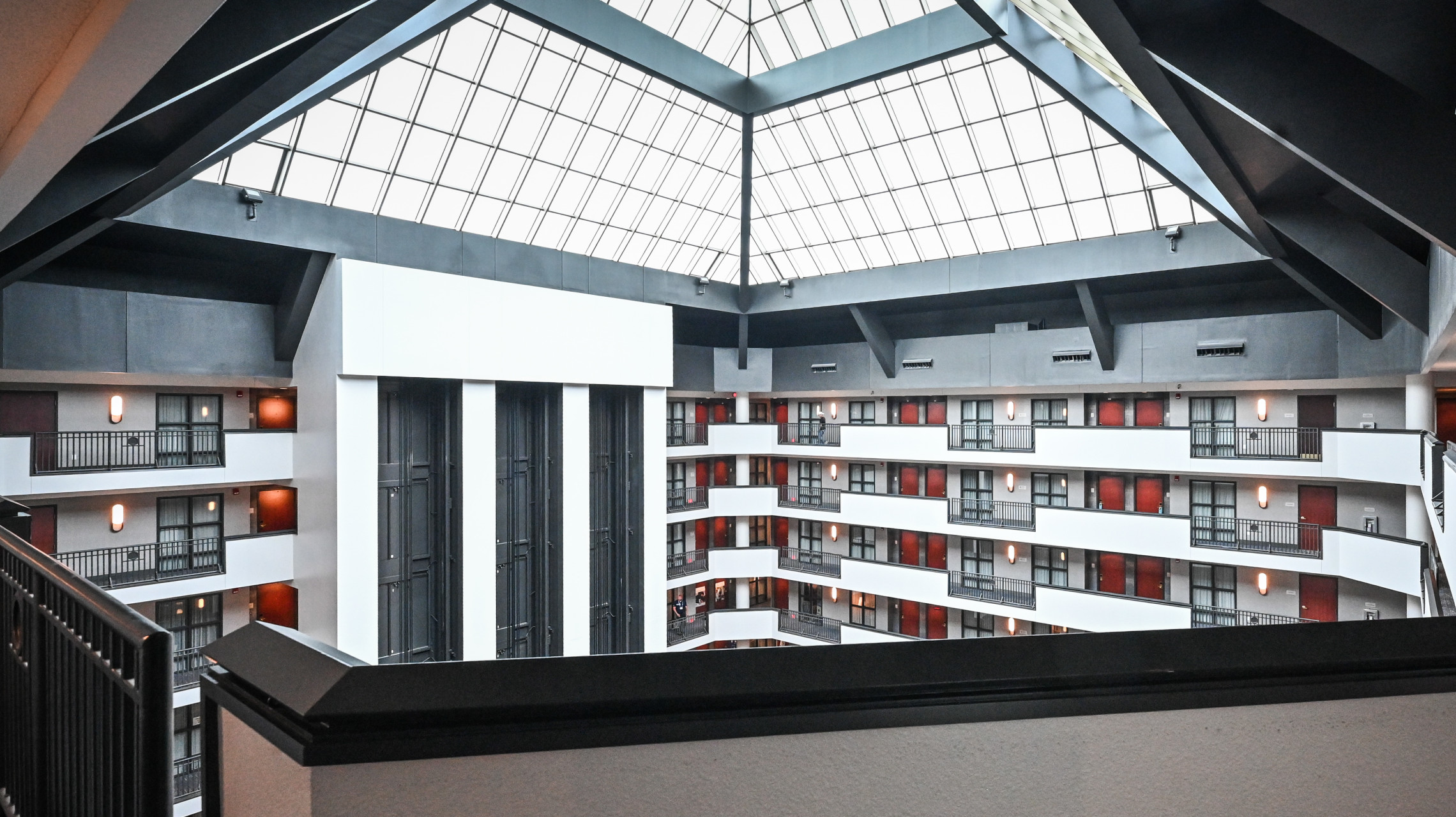 Photograph of a modern multi-story atrium inside a building featuring a large glass skylight ceiling that allows natural light to illuminate the space. The atrium includes multiple floors with white walls, black railings, red doors, and elevator shafts visible on one side.