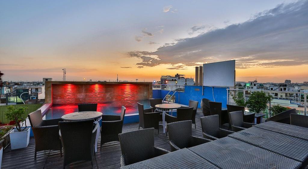 A rooftop terrace during sunset with wicker furniture, a lit pool and a view of the city skyline - TGI Apple Inn, Jaipur 