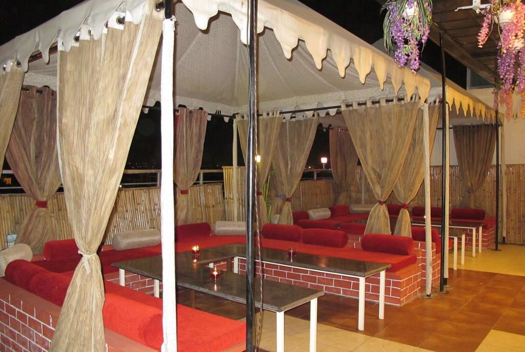 An open-air cabana with sheer curtains, red cushions on bench seating and a view of the city at night - TGI Apple Inn, Jaipur 