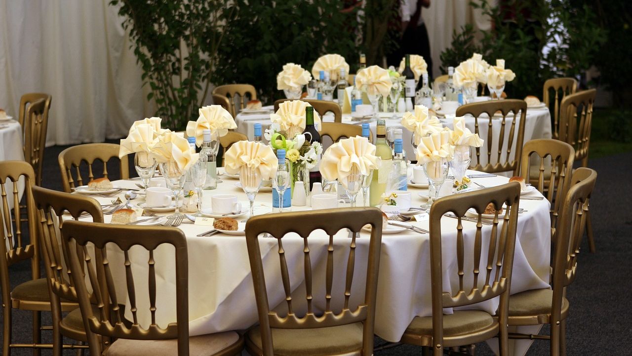 Round banquet tables with white cloths, golden chairs, and cream napkins, adorned with small floral centerpieces and set with dinnerware.