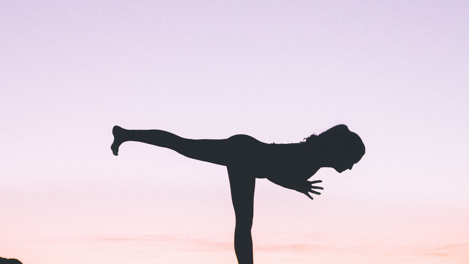 a silhouette of a woman doing a yoga pose amidst nature with pink and purple hued sky in the background