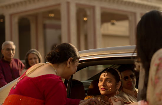 alt-text A joyful scene of a woman arriving in a car greeted by two other women and onlookers; they appear engaged in a warm, celebratory interaction, possibly a cultural event.