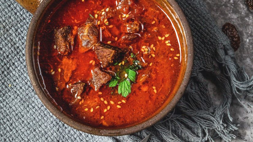 A bowl of red meat curry as seen from above with a fork and a knife kept next to it