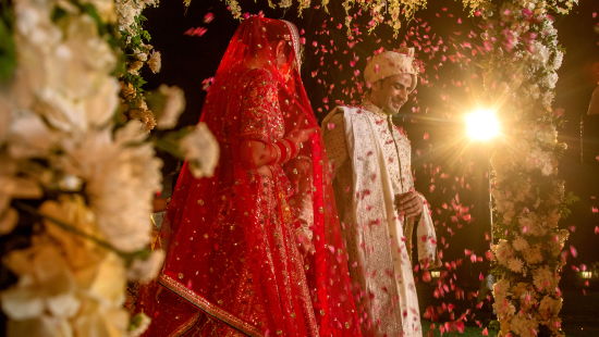 alt-text A bride in a red lehenga stands under a beautifully decorated floral canopy, her face covered with a traditional veil, surrounded by lush flowers and soft lighting.