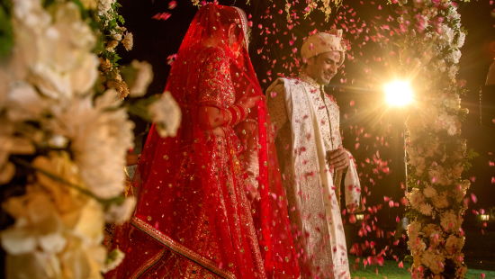 alt-text A bride in a red lehenga stands under a beautifully decorated floral canopy, her face covered with a traditional veil, surrounded by lush flowers and soft lighting.