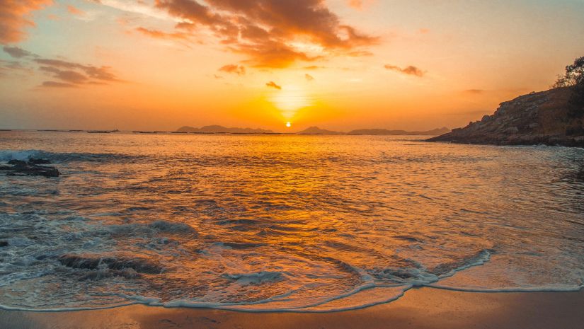 sun setting in the distance and the skies showing shades of blue and orange while the waves crash into the sand