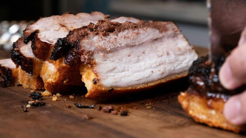 A image of pork roast being cut into pieces with a butcher knife on a wooden table 