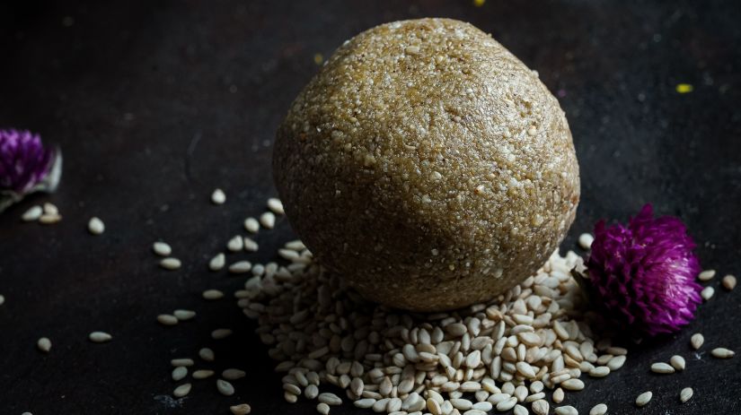 a laddu placed on a plate with sesame seeds below it and flowers next to it