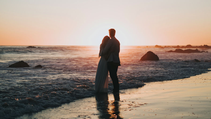 a couple hugging while standing and looking at the sun set on a beach while overlapping waves hits their feet