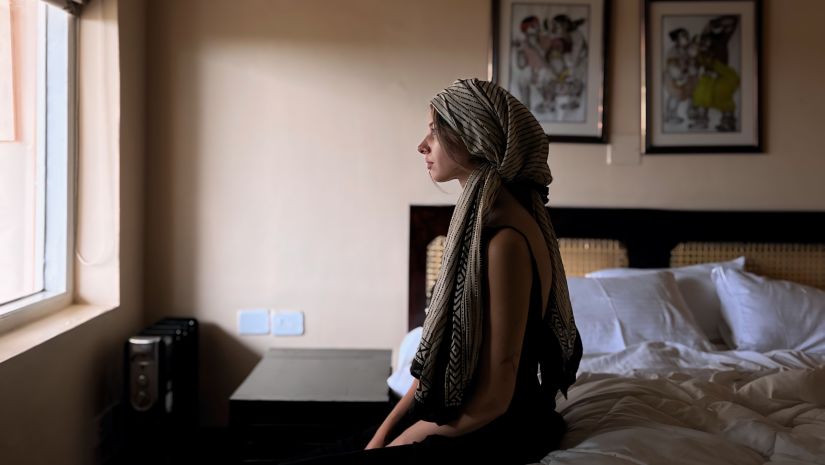 A seated woman wearing a headscarf, looking out a window in a room with a bed and artwork.